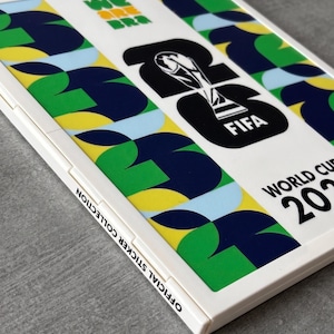 May include: A white rectangular FIFA World Cup 2022 official sticker collection album. The cover features a geometric pattern in green, yellow, blue, and light blue, with the FIFA World Cup trophy icon and the text WE ARE BRA and 2022.