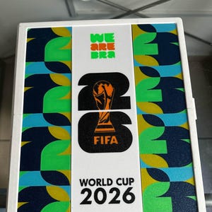 May include: A rectangular white display case featuring the FIFA World Cup 2026 logo in black and orange. The center panel displays the trophy icon, while the side panels feature a repeating geometric pattern in shades of green, blue, and yellow.
