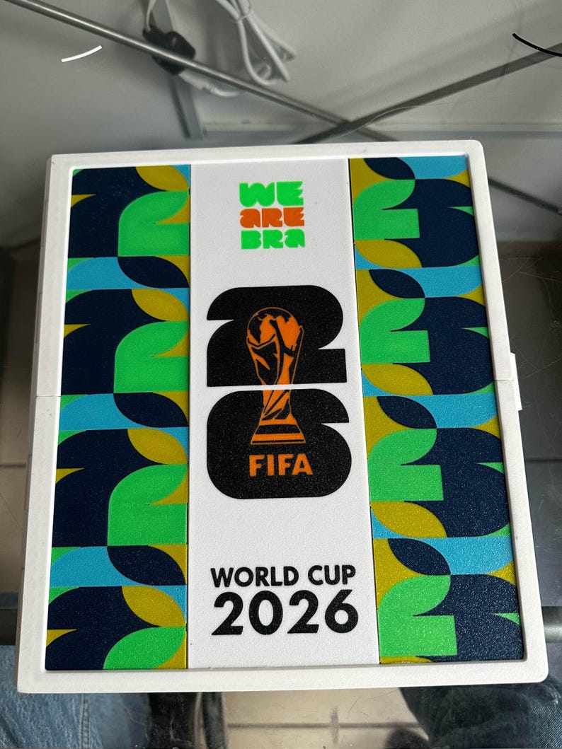 May include: A rectangular white display case featuring the FIFA World Cup 2026 logo in black and orange. The center panel displays the trophy icon, while the side panels feature a repeating geometric pattern in shades of green, blue, and yellow.