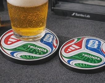 May include: Two round drink coasters featuring a colorful soccer ball pattern in red, green, and blue with the text TRIONDA and the FIFA logo. One coaster supports a glass filled with a golden beverage, while the other sits empty beside it.