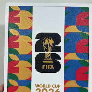 May include: A square white plaque featuring the FIFA World Cup 2026 logo in black with a gold trophy icon. The design includes vertical side panels with a repeating geometric pattern in red, yellow, blue, and green. The text reads FIFA World Cup 2026.