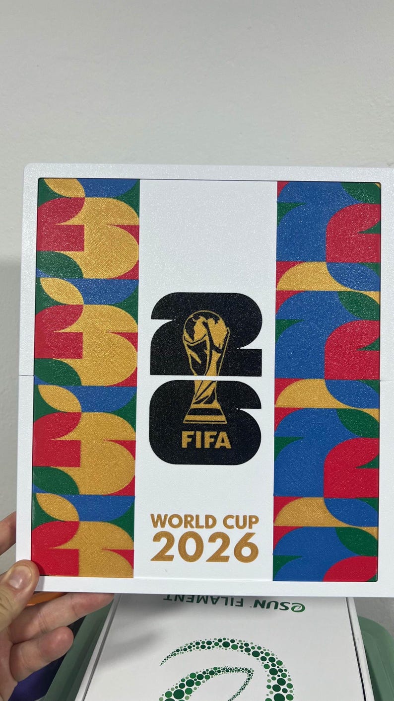 May include: A square white plaque featuring the FIFA World Cup 2026 logo in black with a gold trophy icon. The design includes vertical side panels with a repeating geometric pattern in red, yellow, blue, and green. The text reads FIFA World Cup 2026.