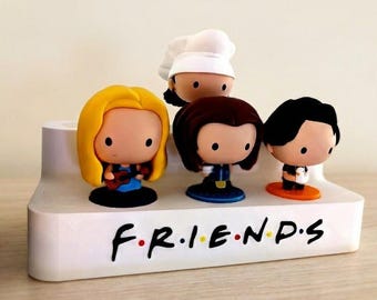 Può includere: Un espositore bianco con la scritta "F.R.I.E.N.D.S" in lettere nere e colorate, che contiene quattro figurine di cartoni animati. Le figurine hanno acconciature e abiti diversi. Una figurina indossa un cappello da chef.
