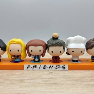 May include: Six Friends character figurines on an orange base. Each figure has a distinct hairstyle and outfit. The base features the word "FRIENDS" in black lettering.