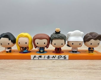 Può includere: Un set di sei statuette dei personaggi di Friends esposte su una base arancione. Ogni personaggio ha un'acconciatura e un abbigliamento unici. La base presenta la scritta "FRIENDS" in lettere nere.