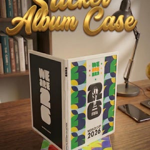 May include: A white FIFA World Cup 2026 sticker album case featuring a black cover with white text reading WE ARE 26 and a colorful cover with the FIFA World Cup 2026 logo, green and blue leaf patterns, and the text WE ARE 26 and WORLD CUP 2026.