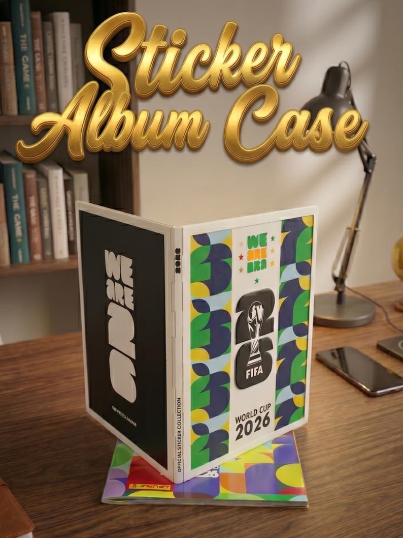 May include: A white FIFA World Cup 2026 sticker album case featuring a black cover with white text reading WE ARE 26 and a colorful cover with the FIFA World Cup 2026 logo, green and blue leaf patterns, and the text WE ARE 26 and WORLD CUP 2026.