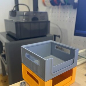 Modular stackable bins on a workbench in a 3D printing studio. The background shows a 3D printer and filament spools, highlighting the product in a real maker workspace.