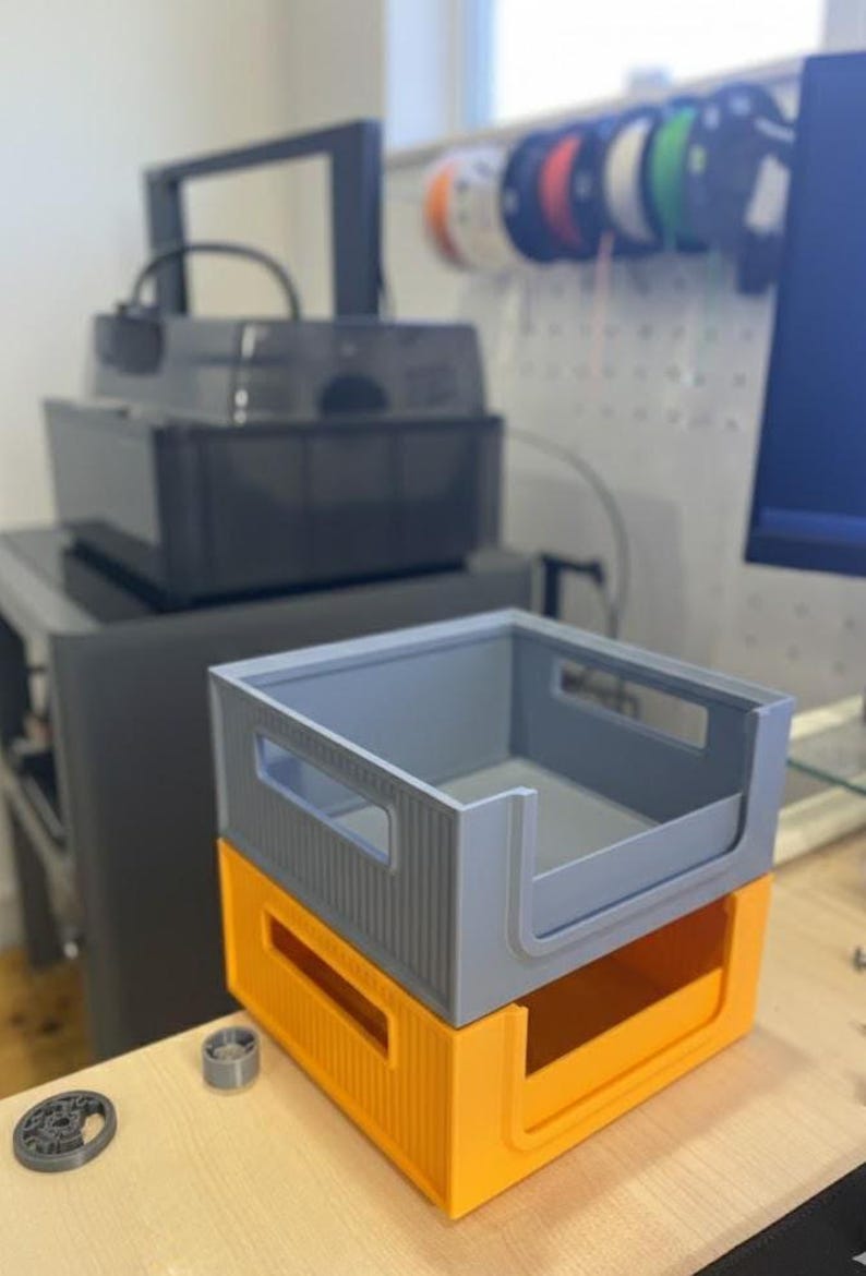 Modular stackable bins on a workbench in a 3D printing studio. The background shows a 3D printer and filament spools, highlighting the product in a real maker workspace.