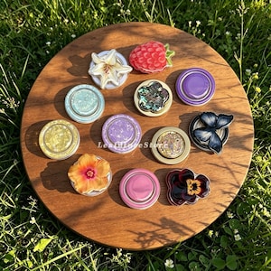 Könnte beinhalten: Eine Sammlung von elf runden Handyhaltern mit verschiedenen Motiven, darunter eine weiße Lilie, eine rote Himbeere, ein blauer Schmetterling, ein gelber Hibiskus, ein dunkles Stiefmütterchen sowie mehrere einfarbige oder texturierte Scheiben.
