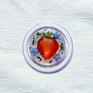 Op de afbeelding: Een transparante harsonderzetter met een geperste rode aardbei en blauwe bloemen. De onderzetter heeft een witte rand en ligt op een witte stoffen achtergrond.