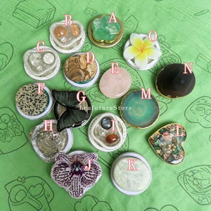 May include: A collection of fifteen circular phone grips labeled A through O, featuring diverse designs including natural stones, floral shapes, a black butterfly, a dalmatian print, and a heart-shaped floral pattern, arranged on a green surface.