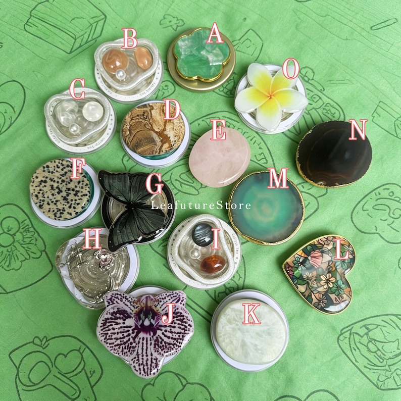 May include: A collection of fifteen circular phone grips labeled A through O, featuring diverse designs including natural stones, floral shapes, a black butterfly, a dalmatian print, and a heart-shaped floral pattern, arranged on a green surface.