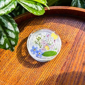 May include: A circular clear resin coaster or paperweight containing pressed botanical elements including small blue forget-me-not flowers, white clusters, yellow petals, thin purple stems, and a single green leaf embedded within the transparent material.