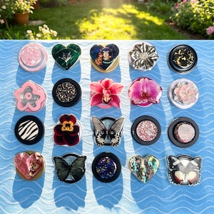 Peut inclure: Une collection de vingt supports de téléphone circulaires et façonnés disposés en grille. Les motifs incluent des fleurs, des papillons, des portraits d'animaux, des paillettes et des textures abstraites dans des tons roses, noirs, dorés et irisés.
