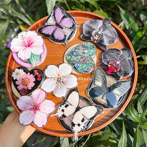 Peut inclure: Une collection de dix supports de téléphone circulaires présentant divers motifs, dont des papillons violets et noirs, des hibiscus et fleurs de cerisier blancs et roses, une incrustation en nacre en forme de cœur et des orchidées sur un plateau en bois.