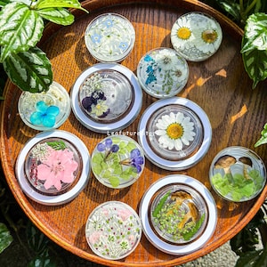 May include: A collection of ten circular clear resin coasters, each embedded with various pressed botanical elements including daisies, blue hydrangeas, pink flowers, purple petals, green ferns, and small mushrooms, arranged on a round wooden tray.