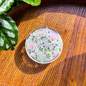 May include: A circular clear phone grip featuring an embedded arrangement of pressed dried flowers in shades of pink, white, and green. The botanical design includes small white blossoms, pink petals, and green foliage encased within the transparent material.