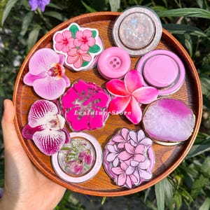 Pode incluir: Uma coleção de dez suportes circulares para telemóvel dispostos numa bandeja redonda de madeira. Os designs incluem flores de hibisco cor-de-rosa, orquídeas roxas e brancas, um botão rosa sólido, resina com flores prensadas e ágata rosa.