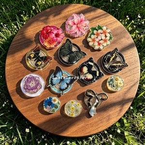 Pode incluir: Uma coleção de suportes circulares para telemóvel com vários designs, incluindo flores de hibisco coloridas, um lírio rosa, plumeria branca, várias borboletas em preto, azul e amarelo, uma flor roxa e pequenos discos de resina com flores silvestres.