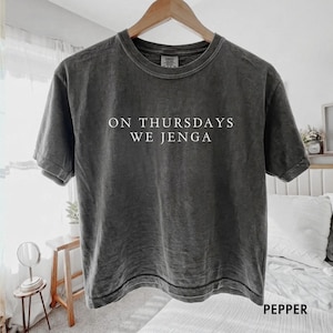 May include: A charcoal gray short-sleeve crew neck t-shirt with a washed-out finish. The center of the chest features white text that reads ON THURSDAYS WE JENGA. The shirt is displayed on a wooden hanger against a light-colored background.
