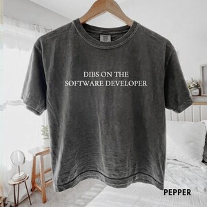 May include: A charcoal grey short-sleeve t-shirt with a vintage-washed finish. The center of the chest features the white text "DIBS ON THE SOFTWARE DEVELOPER" in a clean, sans-serif font. The shirt is displayed on a wooden hanger.