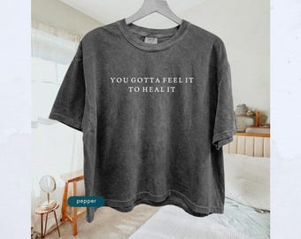 May include: A dark gray, short-sleeved t-shirt with the phrase "YOU GOTTA FEEL IT TO HEAL IT" printed in white. The shirt is hanging on a wooden hanger against a neutral background.