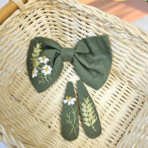 May include: A set of olive green hair accessories, including a bow and two barrettes, all embroidered with white daisies and green leaves. The accessories are displayed on a woven basket.
