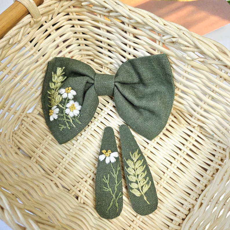 May include: A set of olive green hair accessories, including a bow and two barrettes, all embroidered with white daisies and green leaves. The accessories are displayed on a woven basket.