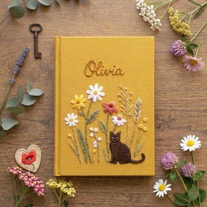 Op de afbeelding: Een mosterdgeel met stof bekleed dagboek met een geborduurd ontwerp van een bruine kat die tussen diverse wilde bloemen en grassen zit. De naam Olivia is in cursief schrift boven het bloemstuk op de voorkant van de kaft geborduurd.