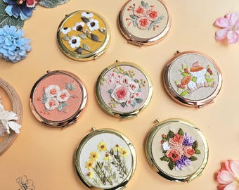 May include: A collection of round, compact mirrors with embroidered floral designs. Each mirror features a different flower arrangement in various colors, set against a neutral fabric background, with gold or rose gold metal frames. The mirrors are arranged on a light orange surface.
