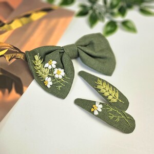 May include: Olive green bow tie and two matching hair clips. Each piece features embroidered white daisies with yellow centers and green leaves. The bow tie and clips are made of a textured fabric.
