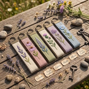 Könnte beinhalten: Sechs rechteckige Stoffschlüsselanhänger mit gestickten botanischen Motiven von Lavendel, Sonnenhut, Lupine, Distel, Lauch und Kornrade, jeweils mit einem bronzefarbenen Metallring und passenden Etiketten auf einer Holzoberfläche.
