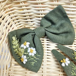 May include: A green linen bow tie and two matching hair clips. The bow tie and clips are embroidered with white and yellow daisy-like flowers and green leaves. The accessories are set against a woven basket background.