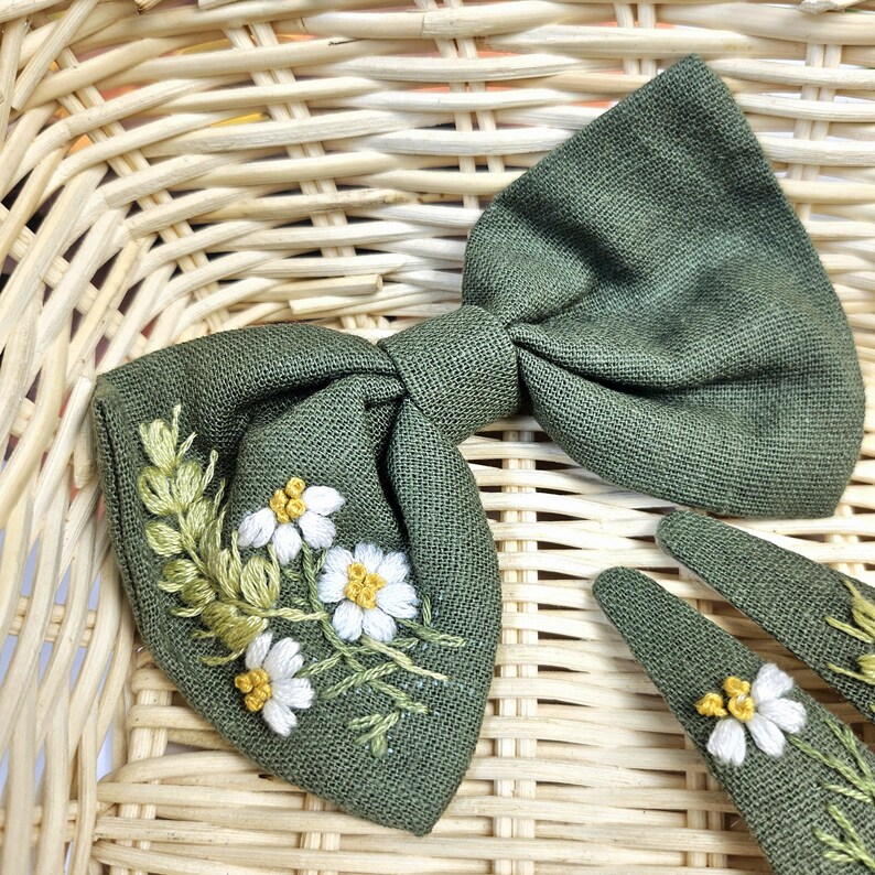 May include: A green linen bow tie and two matching hair clips. The bow tie and clips are embroidered with white and yellow daisy-like flowers and green leaves. The accessories are set against a woven basket background.