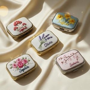 Peut inclure: Cinq petits piluliers rectangulaires avec des motifs brodés. Les boîtes présentent des motifs floraux et de champignons, avec des noms comme "Sophia" et "Sakura" brodés dessus. Les boîtes ont des cadres en métal doré et sont posées sur un tissu crème.