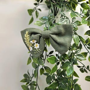 May include: A sage green bow tie with embroidered floral details. The bow tie features a cluster of white daisies and yellow accents on one side. The bow tie is surrounded by green foliage.