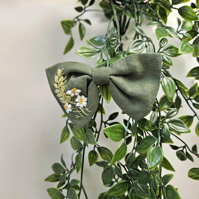 May include: A sage green bow tie with embroidered floral details. The bow tie features a cluster of white daisies and yellow accents on one side. The bow tie is surrounded by green foliage.
