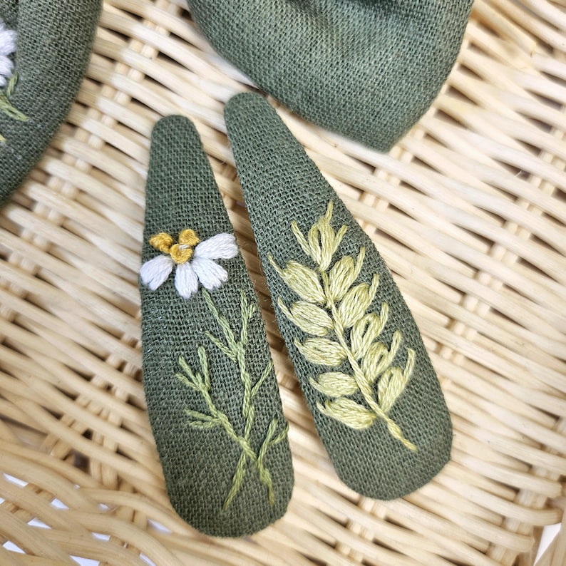May include: Two green fabric hair clips with embroidered floral designs. One clip features a white daisy with a yellow center and green stems. The other clip has a wheat stalk design in shades of yellow and green.