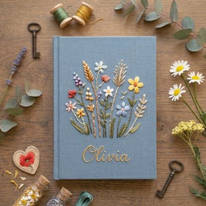 May include: A light blue fabric-covered journal featuring an embroidered bouquet of colorful wildflowers and wheat stalks. The name Olivia is stitched in gold thread below the floral design. The book is rectangular with a hard cover and spine.