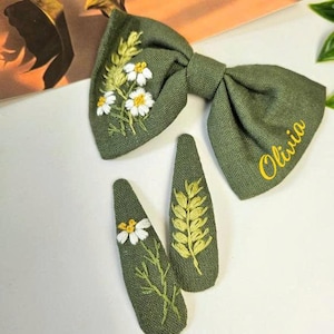May include: Olive green hair bow and two matching barrettes. The bow and barrettes are embroidered with white daisies and yellow accents. The bow has the name "Olivia" embroidered in yellow.
