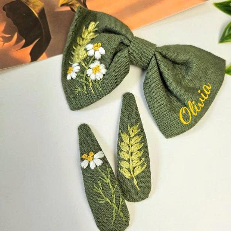 May include: Olive green hair bow and two matching barrettes. The bow and barrettes are embroidered with white daisies and yellow accents. The bow has the name "Olivia" embroidered in yellow.