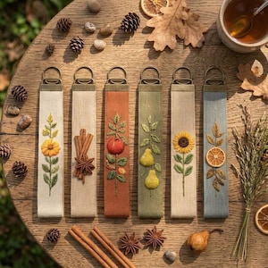 Puede incluir: Seis llaveros de tela rectangulares con motivos otoñales bordados, incluyendo un girasol, ramas de canela, una manzana, peras, un segundo girasol y una rodaja de naranja seca. Cada correa tiene una anilla de metal de color bronce.