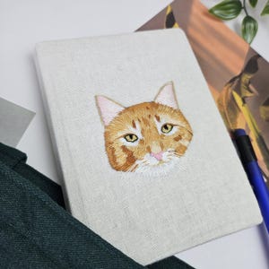 May include: A white linen notebook with a hand-stitched orange tabby cat face on the front cover.
