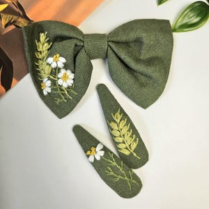 May include: Olive green bow tie and two matching hair clips. The bow tie features white and yellow embroidered flowers and greenery. The hair clips have embroidered wheat and flowers. The accessories are made of a textured fabric.