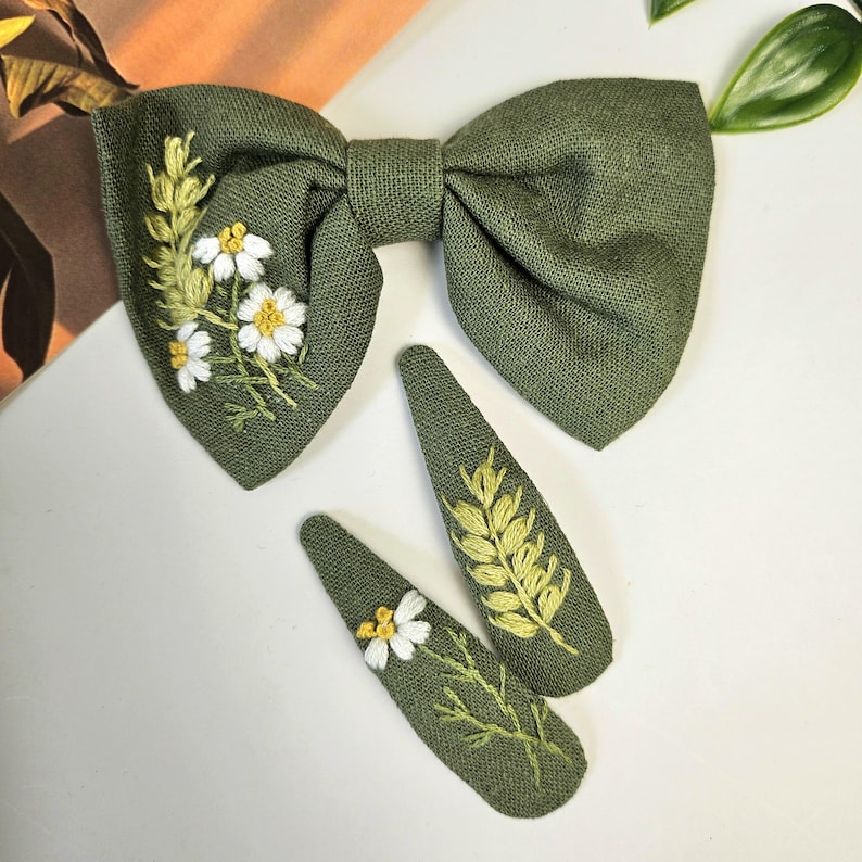 May include: Olive green bow tie and two matching hair clips. The bow tie features white and yellow embroidered flowers and greenery. The hair clips have embroidered wheat and flowers. The accessories are made of a textured fabric.
