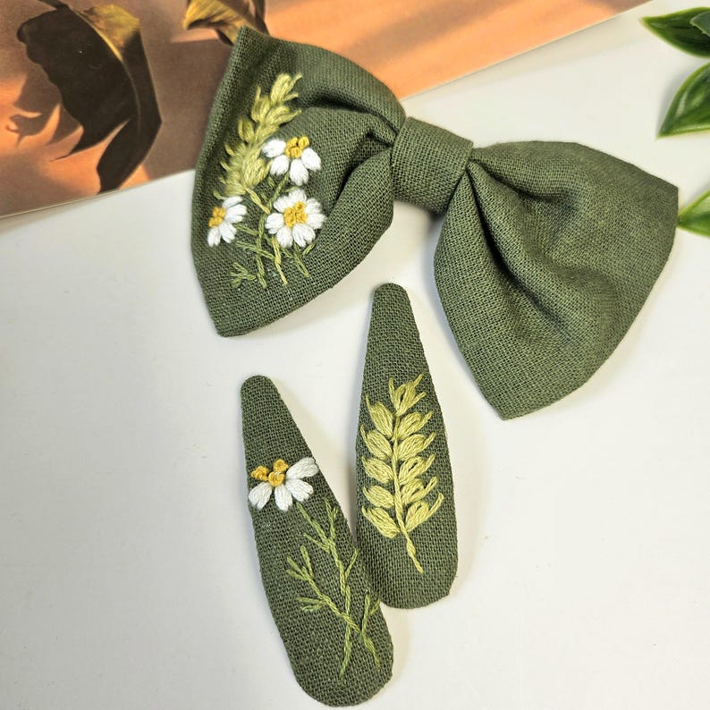 May include: A green linen bow tie and two matching hair clips. The bow tie and clips are embroidered with white daisies and yellow-green leaves. The fabric has a textured appearance.