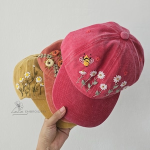 Könnte beinhalten: Drei bestickte Baseballkappen in Gelb-, Orange- und Rottönen. Die Kappen zeigen Blumen- und Bienenmotive. Die Kappen werden in einer Hand vor weißem Hintergrund gehalten. Der Markenname "Lala Embroidery" ist unten links sichtbar.