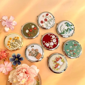 Op de afbeelding: Een verzameling ronde compacte spiegels met geborduurde bloemmotieven. De spiegels hebben zilveren randen en tonen verschillende kleuren, waaronder rood, wit, geel, groen en blauw, met gedetailleerde steken van bloemen en bladeren. De achtergrond is zacht perzik van kleur.
