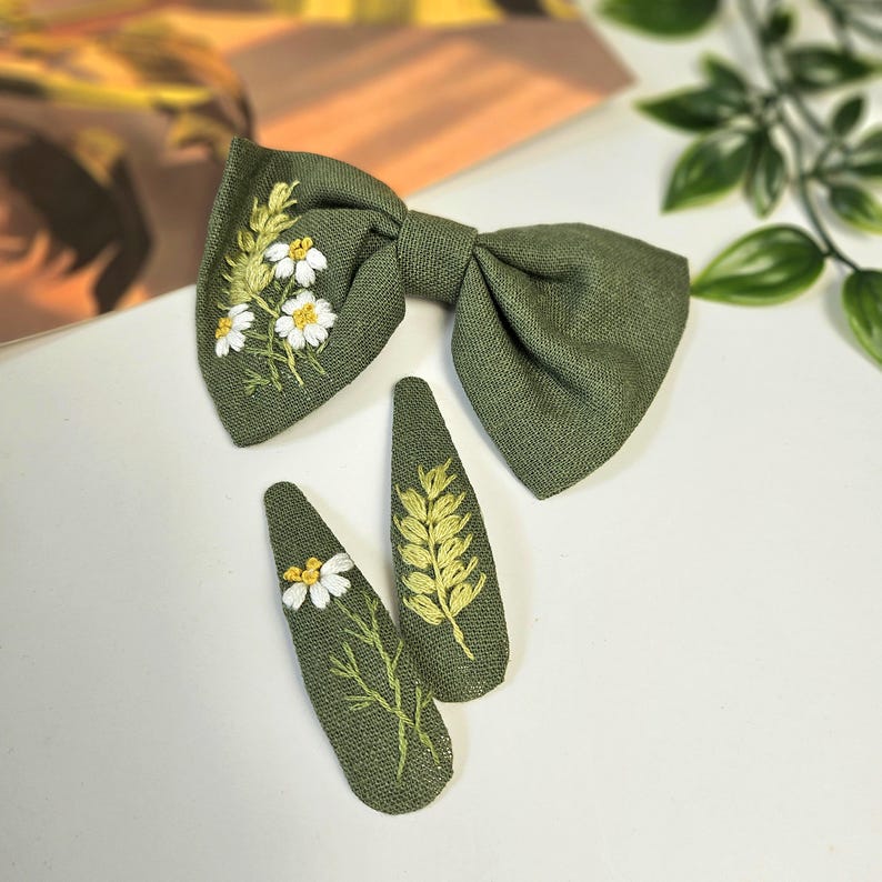 May include: A green linen bow tie and two matching hair clips. The bow tie and clips are embroidered with white daisies and yellow wheat stalks. The accessories are set against a white background.